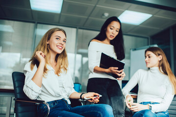 Cheerful Caucasian employee with cellphone technology in hand smiling during collaborative meeting with blurred colleagues, successful business woman holding modern smartphone in office interior