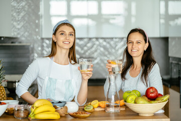 Two different age women, mother and daughter enjoying in kitchen home meal together, one drinking water from glass at hot summer day.