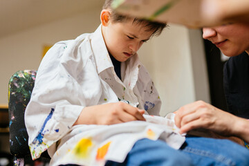 Close up of unrecognizable teacher helping take on white shirt for boy with down syndrome painting...