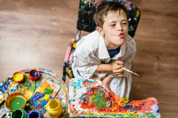 Young Caucasian down syndrome boy sitting at chair with colors and painting on easel with...