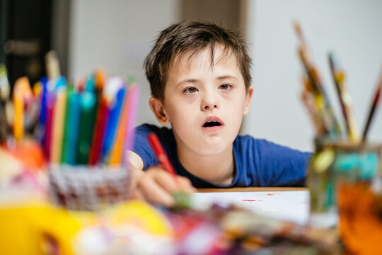 Painting is life for me. Pensive boy with down syndrome looking at camera sitting at an table with white paper while painting with colorful pencil during a painting class.