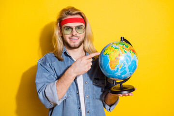 Photo of confident peaceful young gentleman wear jeans shirt dark glasses pointing finger globe smiling isolated yellow color background