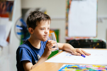 Thoughtful child boy wearing blue t-shirt with down syndrome sitting at table and drinking water from glass at painting studio for special need children.
