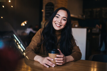Portrait of cheerful Caucasian hipster girl enjoying leisure time in public cafeteria sitting at table and smiling at camera, joyful millennial woman with takeaway cup rejoicing in coffee shop