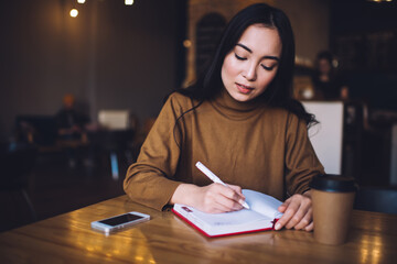 Fototapeta na wymiar Attractive female organisator writing plan ideas in textbook making memo notes with pen, beautiful Caucasian hipster girl spending day in coffee shop for creating publication and learning with notepad