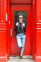 Fashionable Young Woman Wearing Sunglasses, Black Leather Jacket and Bleu Jeans Posing in front of Red Door on Street of New York City.