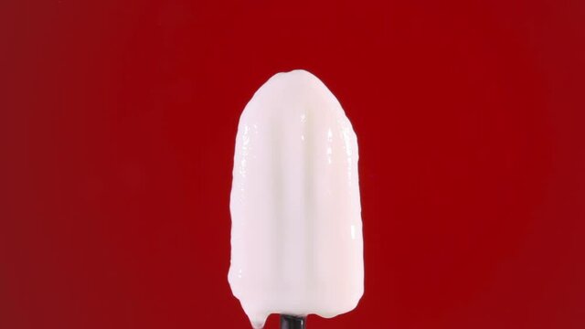 A lemon popsicle (ice pop) melting in a time lapse shot, dissolving; only the black stick remains. Red backdrop.
