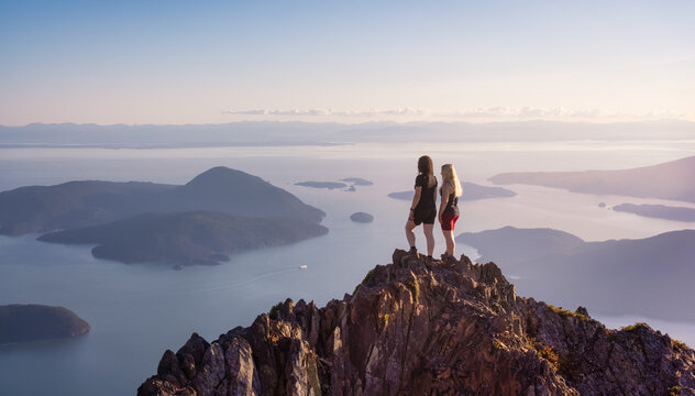 Adventurous Caucasian Woman Friends Hiking on top of a Rocky Mountain Cliff. Sunny Summer Sunset. Mnt Brunswick Hike, North of Vancouver, British Columbia, Canada. - Powered by Adobe
