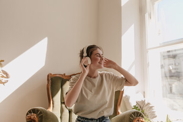 Music, gadgets and chilling concept. Handsome young lady with collected hair, wearing beige t-shirt and white headphones, sitting on armchair at sunlit room near window
