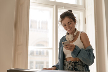 Pretty girl looking at phone in light room. Joyful young lady with collected hair, top, blue shirt and white headphones, smiling and holding mobile