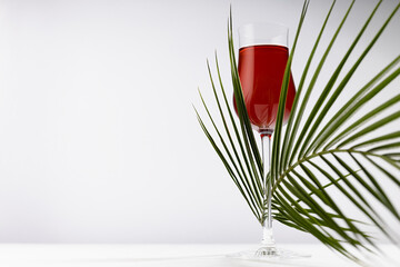 Red liquor in rich glass with elegant curved green palm leaves in sunlight with shadows as...