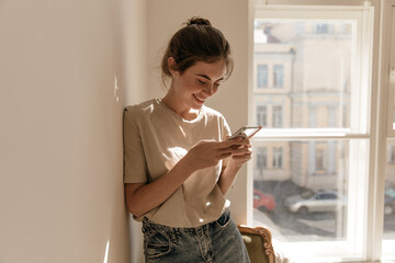Lifestyle portrait of young brunette looking into phone at light room. Pretty girl with collected hair and light clothes, standing against big window background and holding mobile