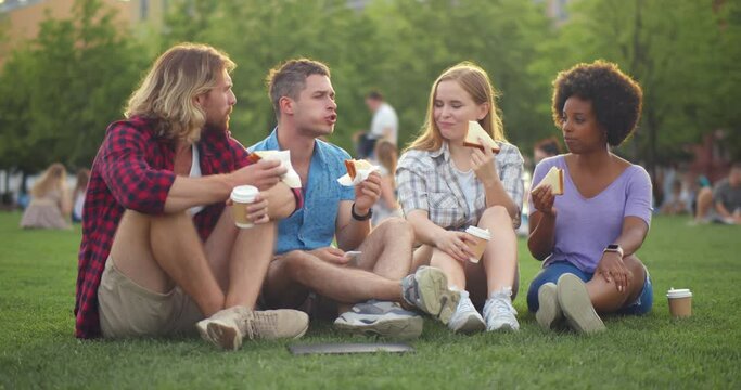 Diverse group of young people sitting on grass and eating sandwiches with coffee during picnic in park