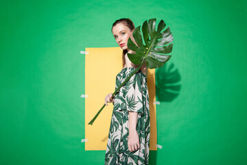 Young female model in stylish dress with floral ornament holding monstera leaf while standing against yellow paper sheet on green background in summer © julenochek