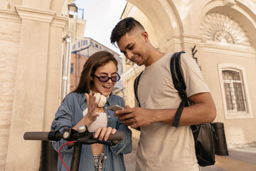 Lifestyle portrait of young joyful couple against beige building background. Excited dark-haired lady in blue shirt, sunglasses, holding electric scooter and looking at phone near friend