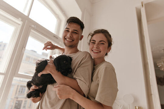 Two friends posing with black little dog. Young girl in loungewear with collected hair, hugging man holding puppy in light room against window background