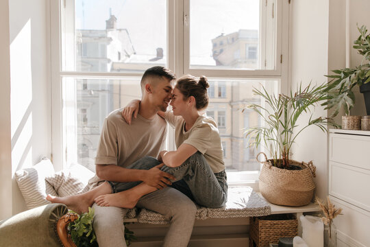 Cute young couple sitting on windowsill against city view background. Pretty girl with brunette collected hair in jeans and t-shirt relaxing near boyfriend in light clothes indoor - Powered by Adobe