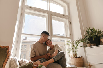 Family and relationship concept. Pretty young boy wearing light loungewear, kissing girlfriend in jeans and t-shirt. Pair sitting on windowsill at home near plants