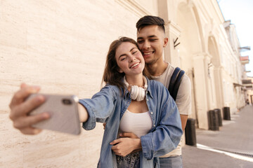 Joyful young couple making selfie-portrait in sunny city. Gorgeous dark-haired man smiling and hugging girlfriend, wearing white top and headphones, blue shirt