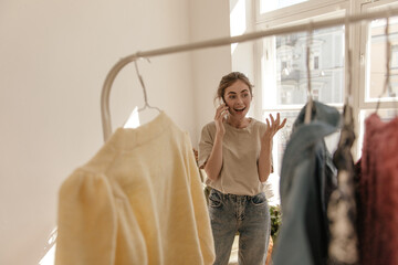 Lifestyle portrait of young lady worrying and talking on phone at light room with clothes. Pretty dark-haired girl having conversation against big window