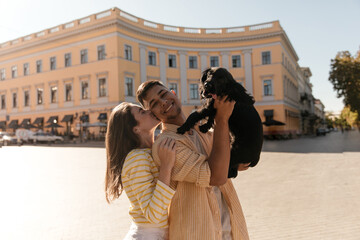 Lifestyle portrait of young couple against city landmark background at sunny warm day. Cute dark-haired girl in yellow outfit kissing and hugging boyfriend, holding little black dog