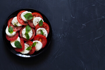 Italian caprese salad with green on the black plate with copy space. sliced red tomatoes, mozzarella cheese, green basil, olive oil on a dark background. fresh salad.top view.