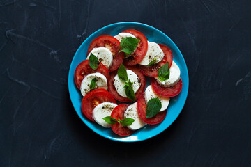 traditional Italian caprese salad on the blue plate. sliced red tomatoes, mozzarella cheese, green basil, olive oil on a black background. fresh salad with copy space. top view.flat lay