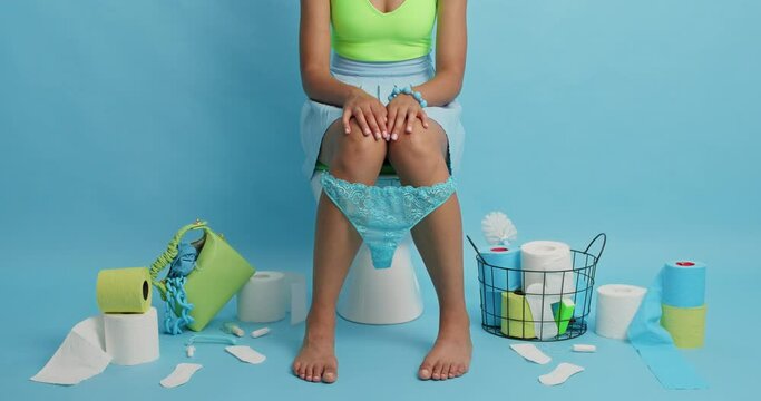 Unknown woman poses on toilet bowl rolls out toilet paper reveals herself wears lacy panties on legs surrounded by hygiene products isolated over blue background. Sanitary and leisure concept