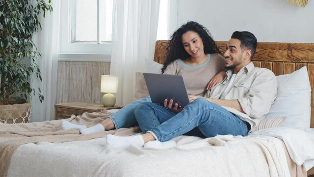 Spaniard arab married couple man and woman lying on bed in cozy bedroom home using laptop for video call making internet purchase in network computer app choosing goods movie smiling looking at screen