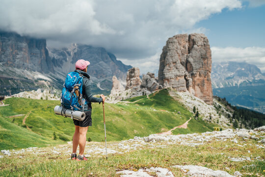 Female trekker walking with backpack and trekking poles by green mountain hill and enjoying the picturesque Dolomite Alps Cinque Torri formation. Active people and mountains concept.