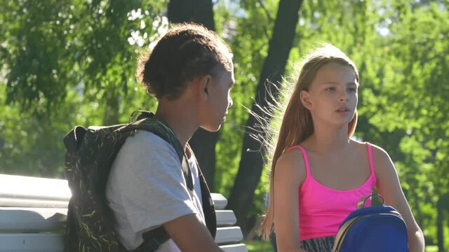 Multicultural teenagers with backpack talking on bench outdoors