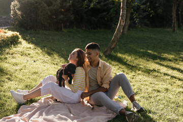 Family, picnic, happiness, relationship concept. Adorable young lady wearing yellow blouse, light pants and sneakers, holding dog and sitting on wrap near husband outdoors at sunny day