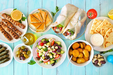 Greek food table scene, overhead view on a blue wood background. Souvlaki, gyros wraps, salad, spanakopita, pita and tzatziki, lemon potatoes, dolmades.