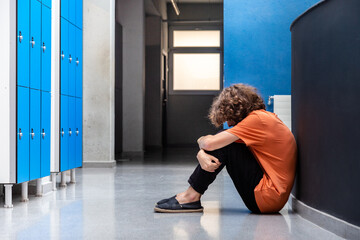 Sad caucasian teen boy victim of bullying sitting on floor in high school corridor. Copy space.
