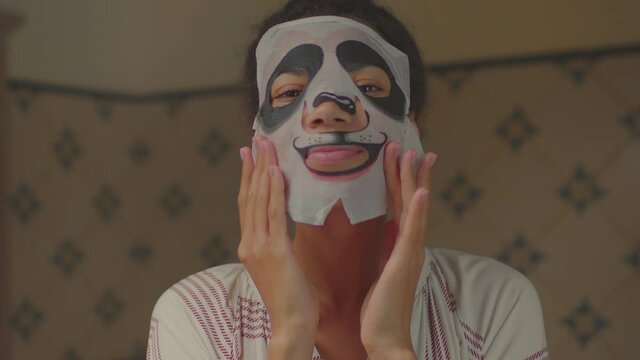 20s African American woman wearing funny panda cosmetics facial mask in bathroom looking at camera. Funny woman enjoying beauty procedures at home.