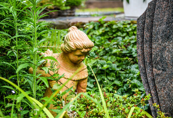 Figur eines kleinen Mädchens neben einem Grabstein inmitten von grünen Pflanzen