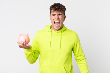 young handsome man shouting aggressively, looking very angry and holding a piggy bank