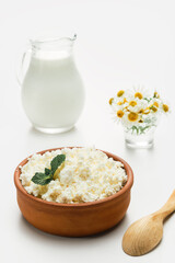 Cottage cheese in a ceramic rustic cup, next to a wooden spoon and a jug of milk. Close-up, selective focus, bright white background. Soft curd , natural healthy food, complete diet food