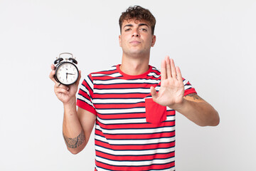 young handsome man looking serious showing open palm making stop gesture and holding an alarm clock