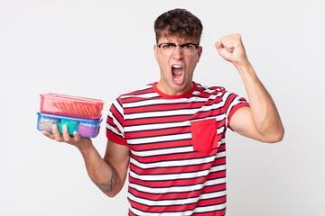 young handsome man shouting aggressively with an angry expression and holding a lunch boxes