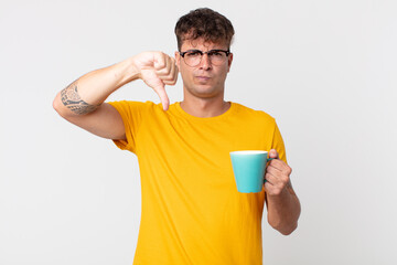 young handsome man feeling cross,showing thumbs down and holding a coffe cup