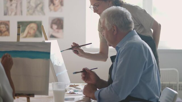 A female teacher shows a retired man how to draw a picture with paints and a brush at courses for the elderly. A senior man draws a picture to a group of pensioners
