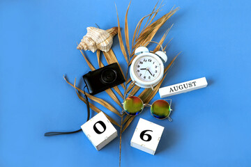 Calendar for August 6 : the name of the month of August in English, cubes with the numbers 0 and 6, a dry palm leaf, a shell, a camera, a watch, sunglasses on a blue background, a top view