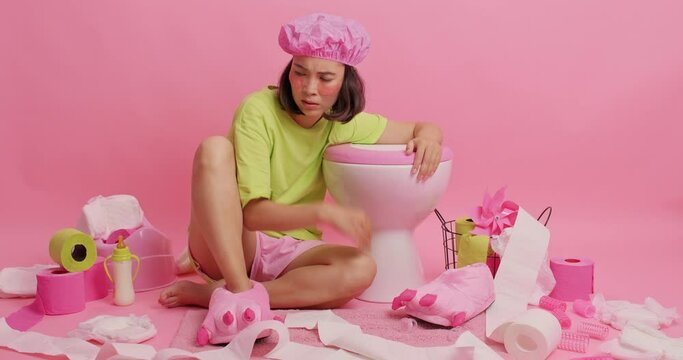 Tired Asian woman gestures angrily poses in restroom near toilet bowl being annoyed with mass wears bath hat casual green t shirt applies beauty patches under eyes isolated over pink background