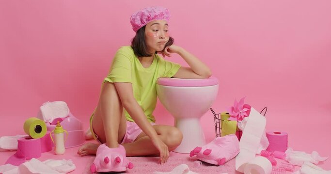 Frustrated Asian woman wears shower cap t shirt and shorts shrugs shoulders leans on toilet bowl applies beauty patches under eyes smiles happily surrounded by different items isolated over pink wall
