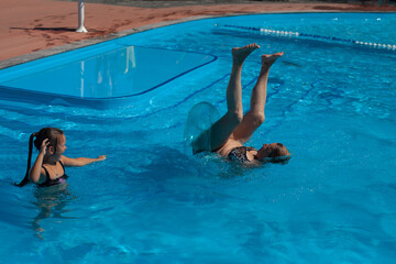 Grandmother and granddaughter in the pool. The grandmother falls upside down from the inflatable circle into the pool water, the granddaughter laughs and smiling. 