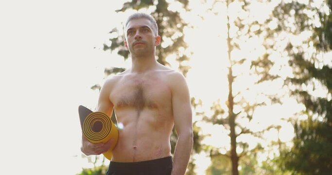 Portrait of Young Man Yog. He Standing in Park with Sport Mat in Hands and Looking forward. Yoga Training in Park. Concept of Healthy Lifestyle. Slow motion