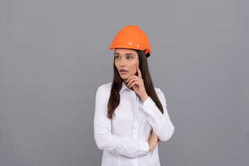 thoughtful woman in safety hardhat and white shirt on grey background, builder