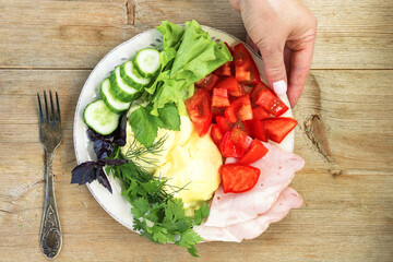 Female hand put omlette with tomato, cucumber, herbs, ham, lettuce on old wooden table with fork. Morning breakfast concept. Close up. Selective soft focus. Text copy space.
