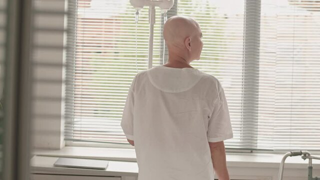 Rear-view medium panning slowmo of bald woman with cancer wearing white gown standing in front of window in hospital chamber looking at catheter in her arm